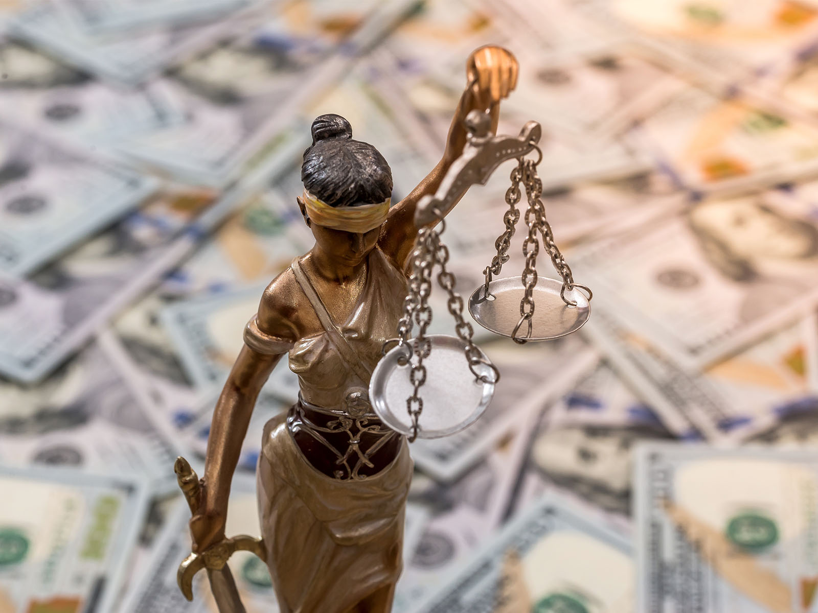 This photo illustration shows a bronze statue of blind Justice holding scales. The background is a scattering of U.S. dollars. This art is related to a story about AML Compliance, money laundering, etc.